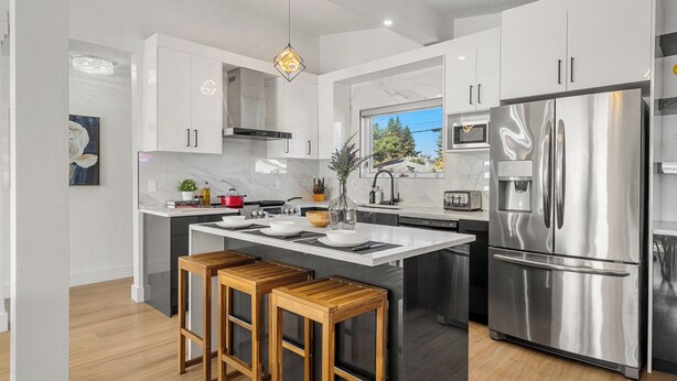 A modern kitchen with white cabinets, a stainless steel refrigerator, wooden bar stools, and a large window. - Perfect for families & groups seeking comfort, games & style in Edmonton (Edmonton)