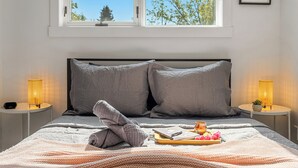 A neatly made bed with a grey quilt, two bedside lamps, a tray with snacks, and a window with a view of trees. - Perfect for families & groups seeking comfort, games & style in Edmonton (Edmonton)