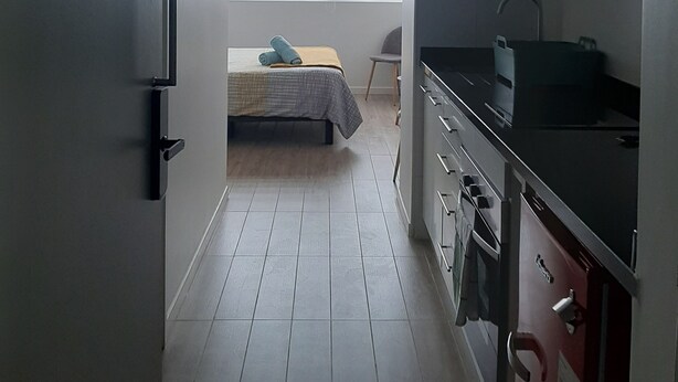 Een compacte keuken met een bed op de achtergrond, een klein eethoekje en een raam met zonwering.