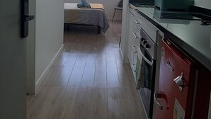 Een kleine keuken met een gootsteen, fornuis en kastjes. In de achtergrond is een bed met een blauw kussen te zien.