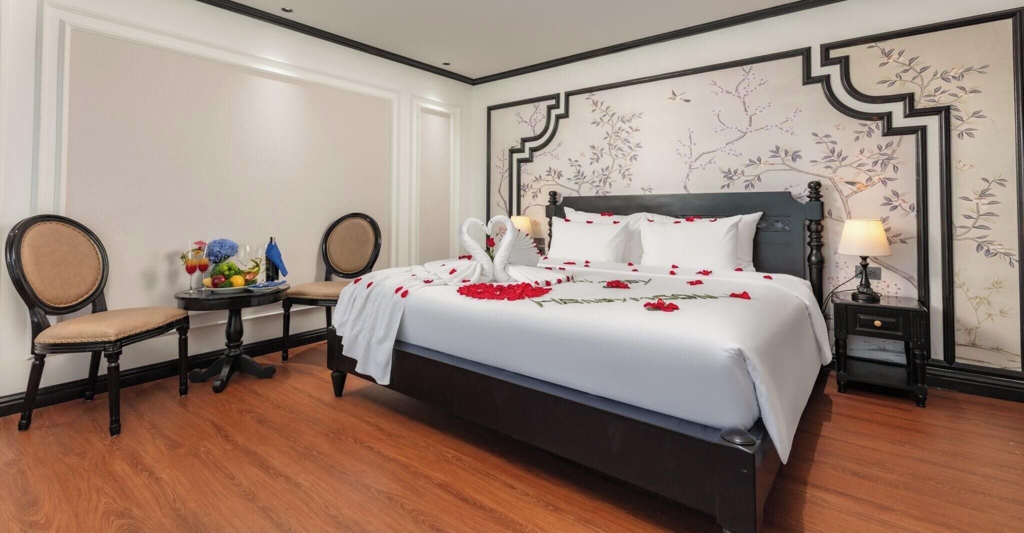 Habitación de hotel con una cama grande, una mesita de noche con lámpara, una silla y una pequeña mesa con fruta y flores.