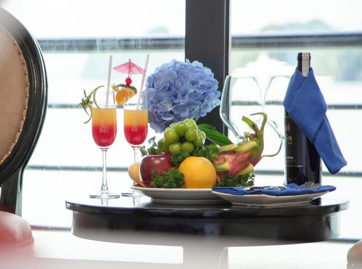 A table with a bottle of champagne, a blue napkin, a fruit bowl, two drinks with straws, and a decorative blue flower.