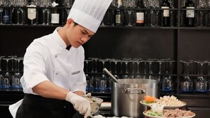 A chef preparing food in a kitchen with wine bottles and glasses on shelves behind. - Halora Nova Cruise (Tuan Chau)