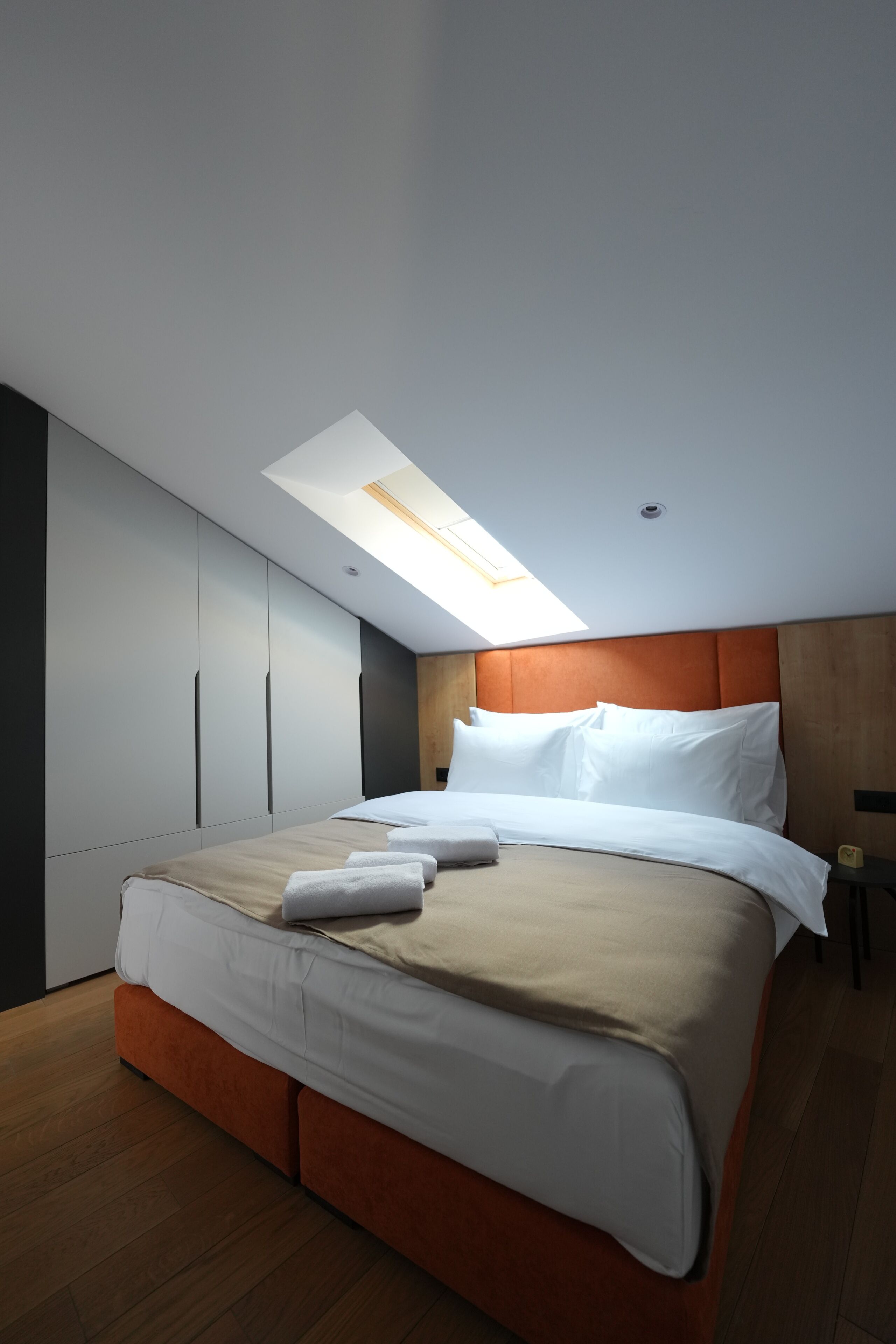 A modern bedroom with a skylight, a bed with white linens, and wooden flooring.