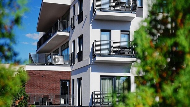 A modern multi-story building with balconies, a brick facade, and a paved entrance area.