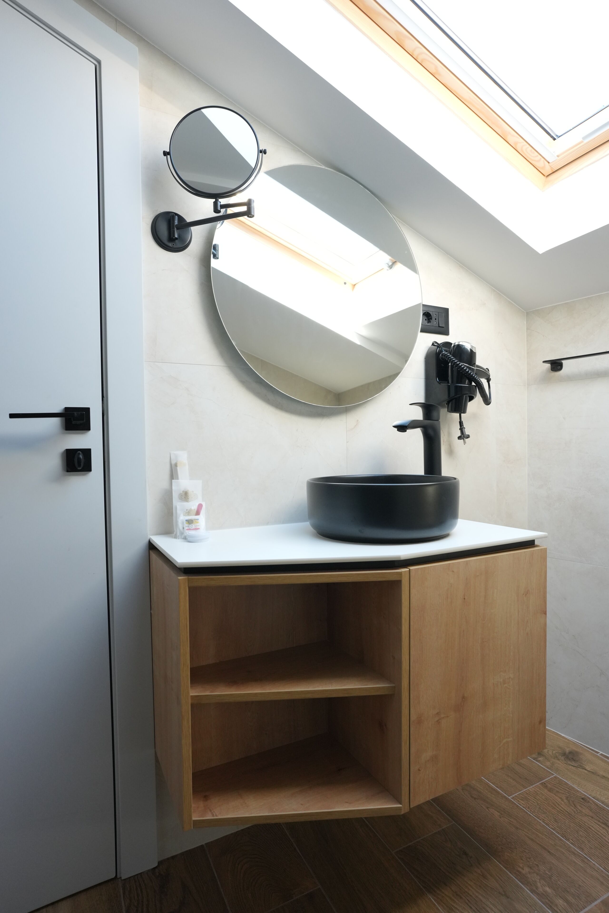 A modern bathroom with a round mirror, black sink, wooden cabinet, and a skylight.
