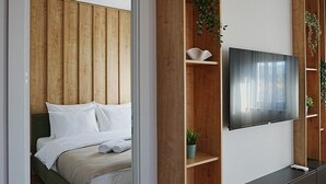 A modern bedroom with a wooden headboard, a bed with white linens, a TV mounted on the wall, and built-in shelving with plants. - Madera (Čačak)