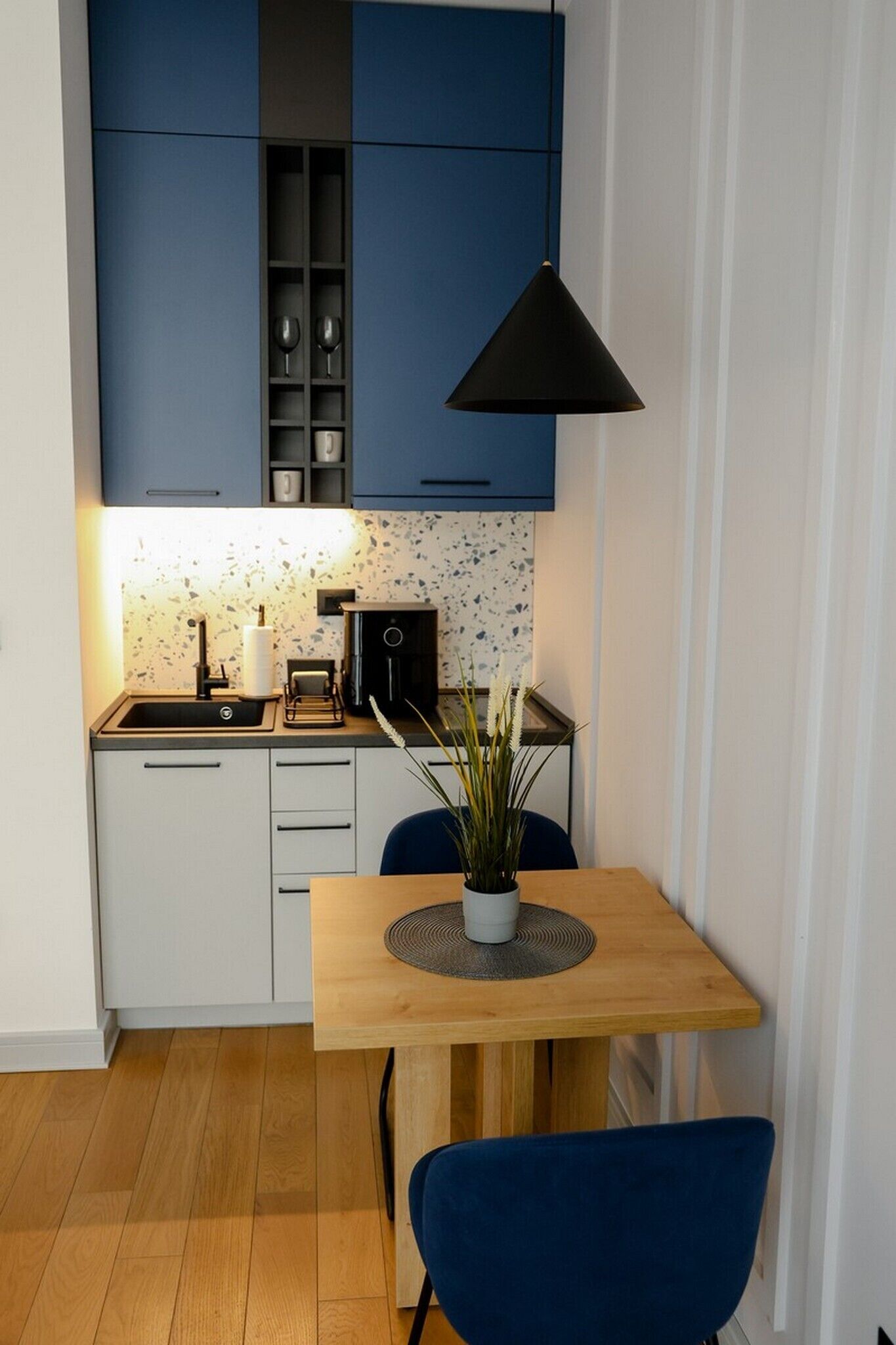A compact kitchen with a sink, stove, and cabinets, a dining table with a chair, and a potted plant.