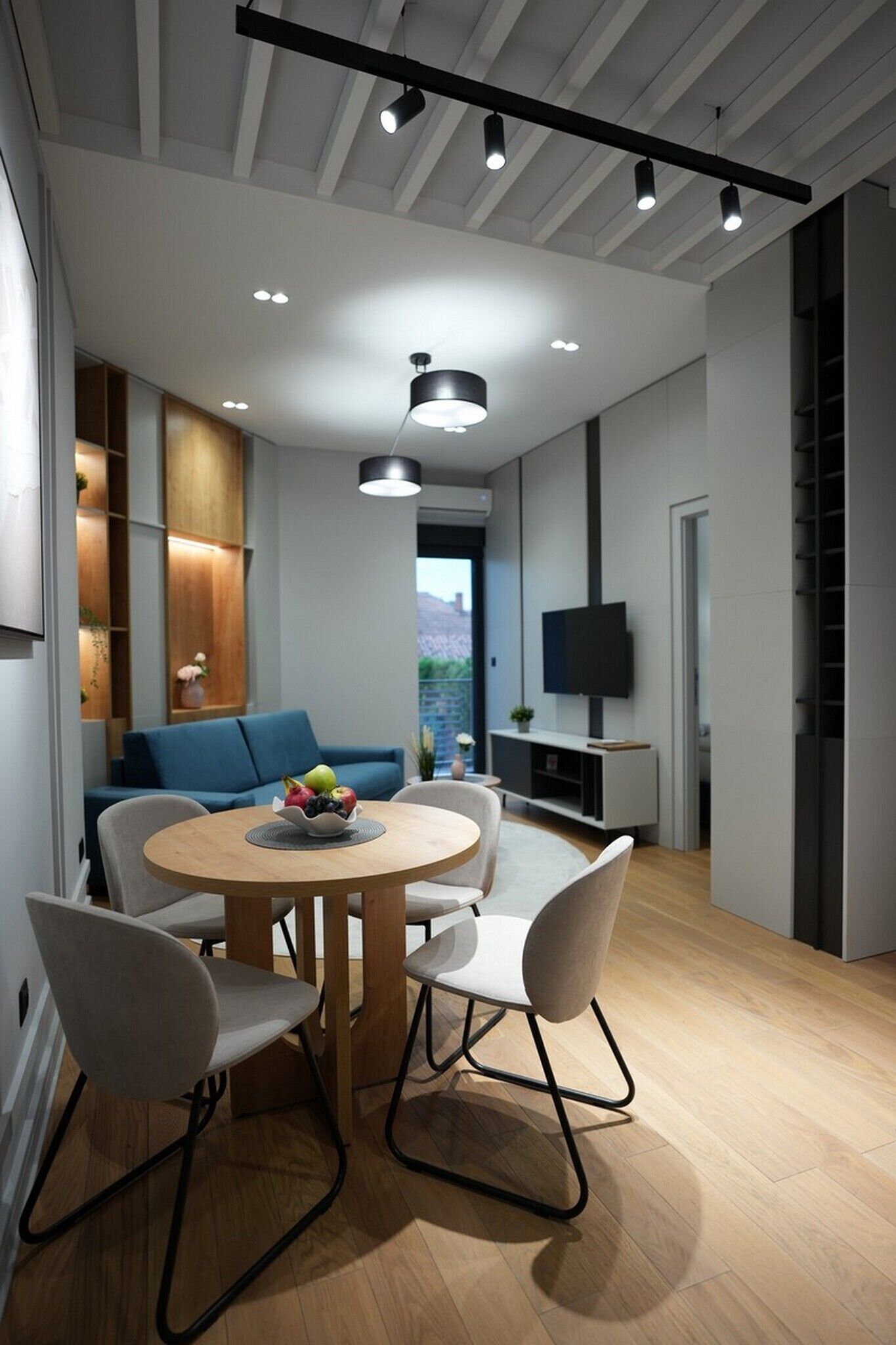 A modern living room with a round wooden dining table, grey upholstered chairs, a blue sofa, and a TV on a white stand.