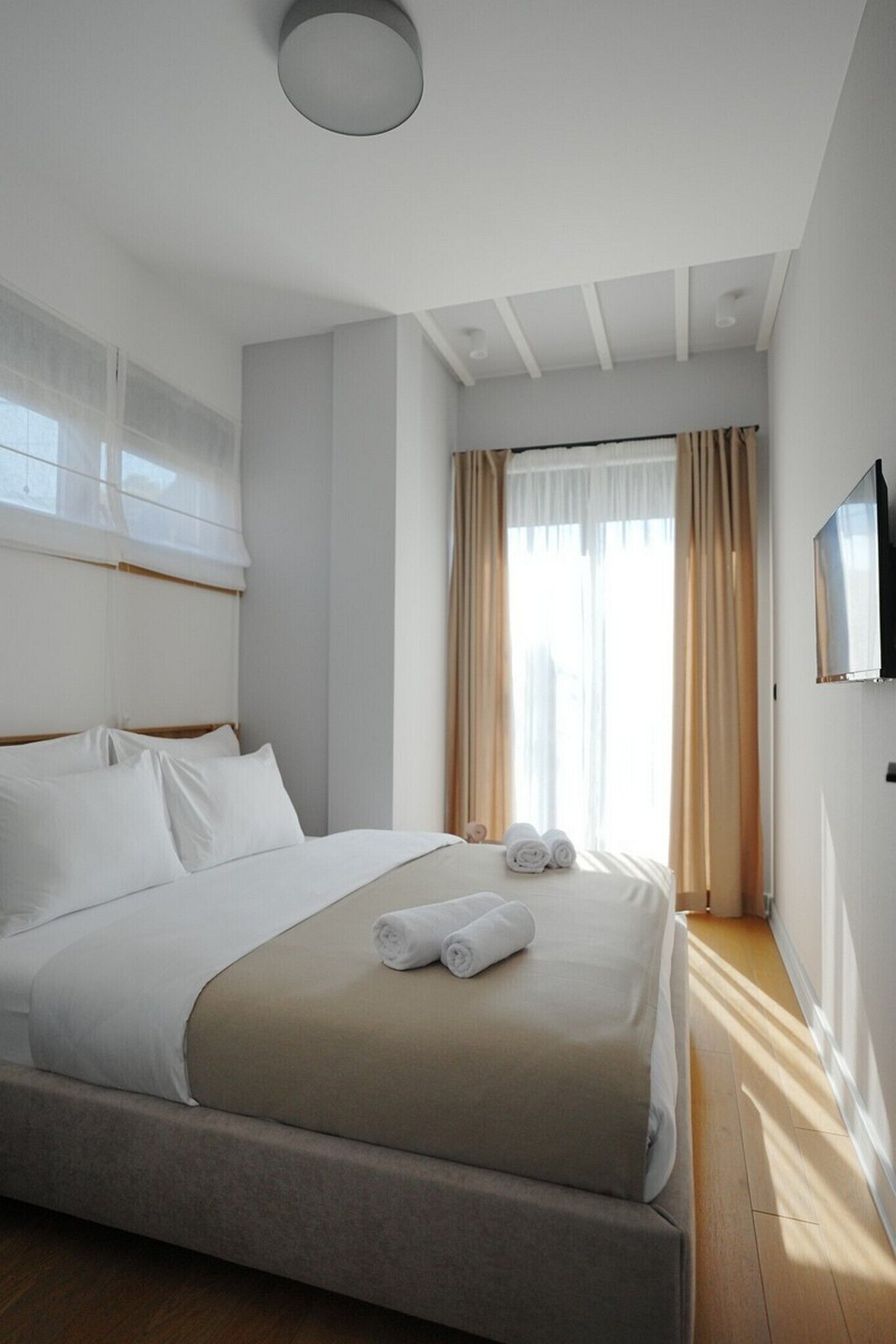 Ein modernes Hotelzimmer mit einem großen Bett, beiger Bettwäsche und Blick auf die Stadt durch transparente Vorhänge.