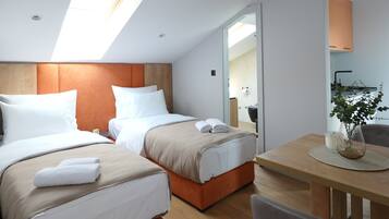 A modern hotel room with two beds, a dining table, and a kitchenette.