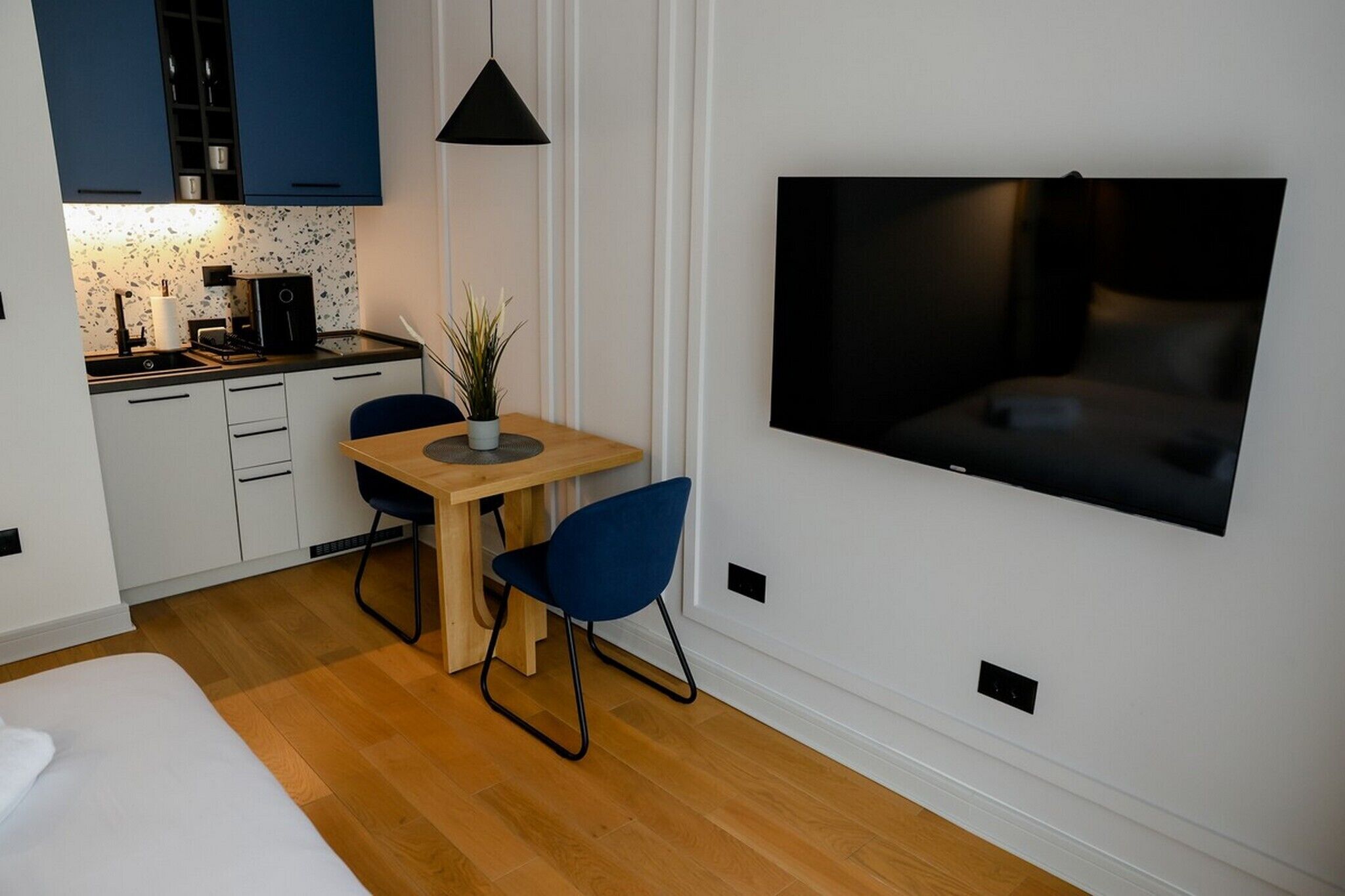 A compact hotel room with a kitchenette, a dining area, a bed, and a TV mounted on the wall.