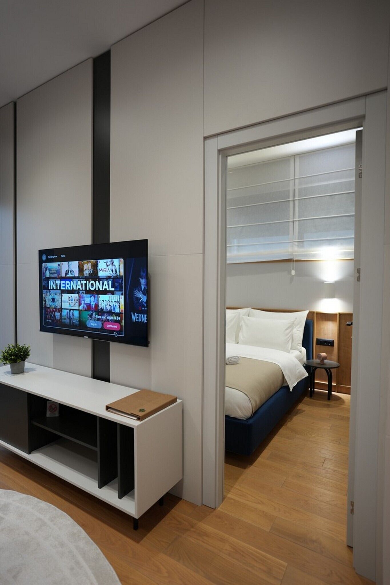 A modern hotel room with a flat-screen TV displaying a news channel, a bed with a blue headboard, a bedside table with a lamp, and a wooden floor.