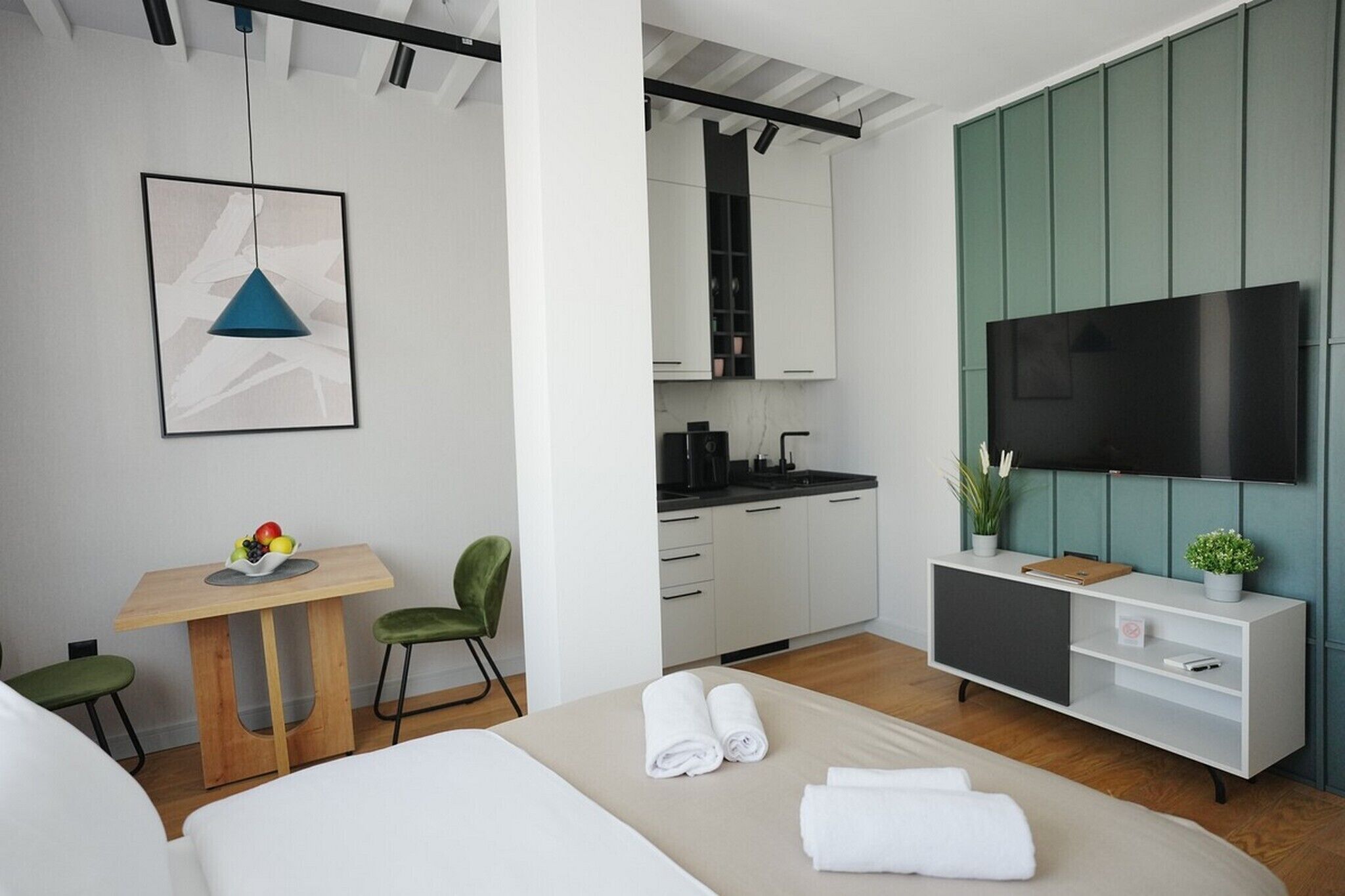Et moderne hotelværelse med seng, skrivebord med stol, et lille tekøkken og et fjernsyn ophængt på væggen.