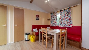 A room with a dining table, chairs, a red sofa, a window with patterned curtains, and a wooden floor. - Mobile Home “Les Volets rouges” with Private Terrace (Plouhinec)