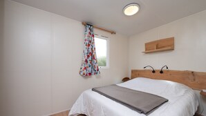 A bedroom with a wooden headboard, a window with a curtain, a bedside table, and a bed with a blanket. - Mobile Home “Les Volets rouges” with Private Terrace (Plouhinec)