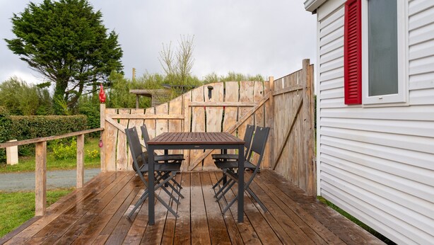 A wooden deck with a table and chairs, a wooden fence, and a white building with red shutters. - Mobile Home “Les Volets rouges” with Private Terrace (Plouhinec)
