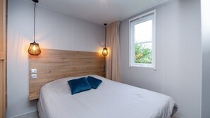 A modern bedroom with a wooden headboard, two bedside lamps, a window with a view, and a bed with a blue pillow. - Mobile Home "Au vert du jardin" 500m from the Beach (Plouhinec)