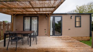 A wooden deck with a table and chairs, a covered patio area, and a sliding glass door leading to a covered outdoor space. - Mobile Home "Au vert du jardin" 500m from the Beach (Plouhinec)