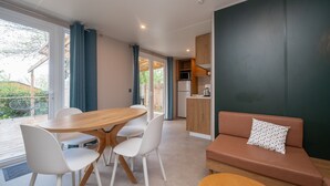 A modern kitchen with a microwave and refrigerator, a wooden dining table with white chairs, a brown sofa with a patterned pillow, and a sliding glass door leading to a patio. - Mobile Home "Au vert du jardin" 500m from the Beach (Plouhinec)