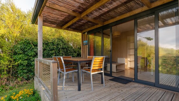 A wooden deck with a table and chairs, a glass door leading to an interior space, and a view of greenery. - Chalet "35 m² - Molène" in Plouhinec, 5 Guests, 2 Bedrooms (Plouhinec)