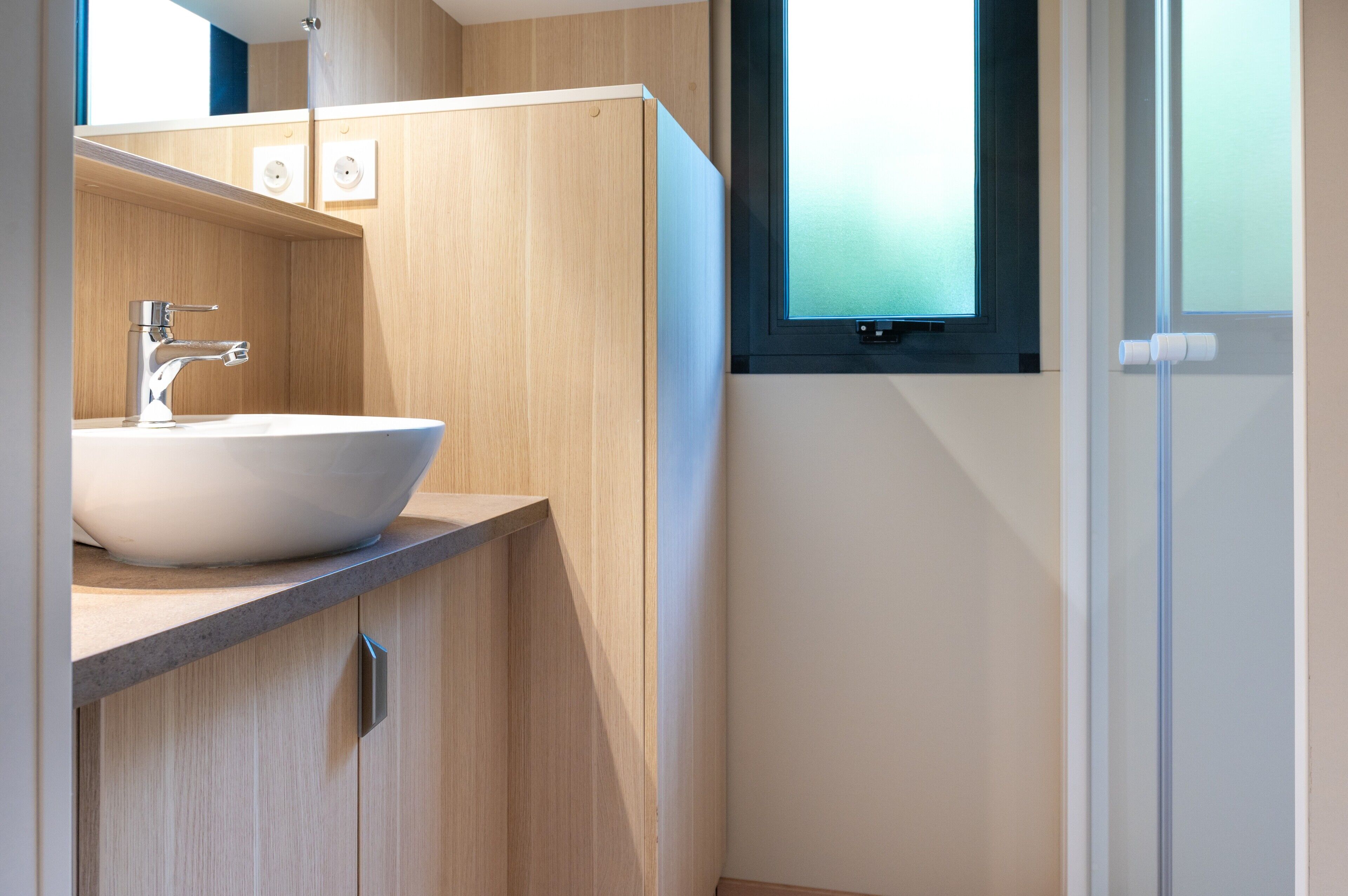 Un bany modern amb un lavabo blanc, armaris de fusta i una finestra amb vidre acidulat.
