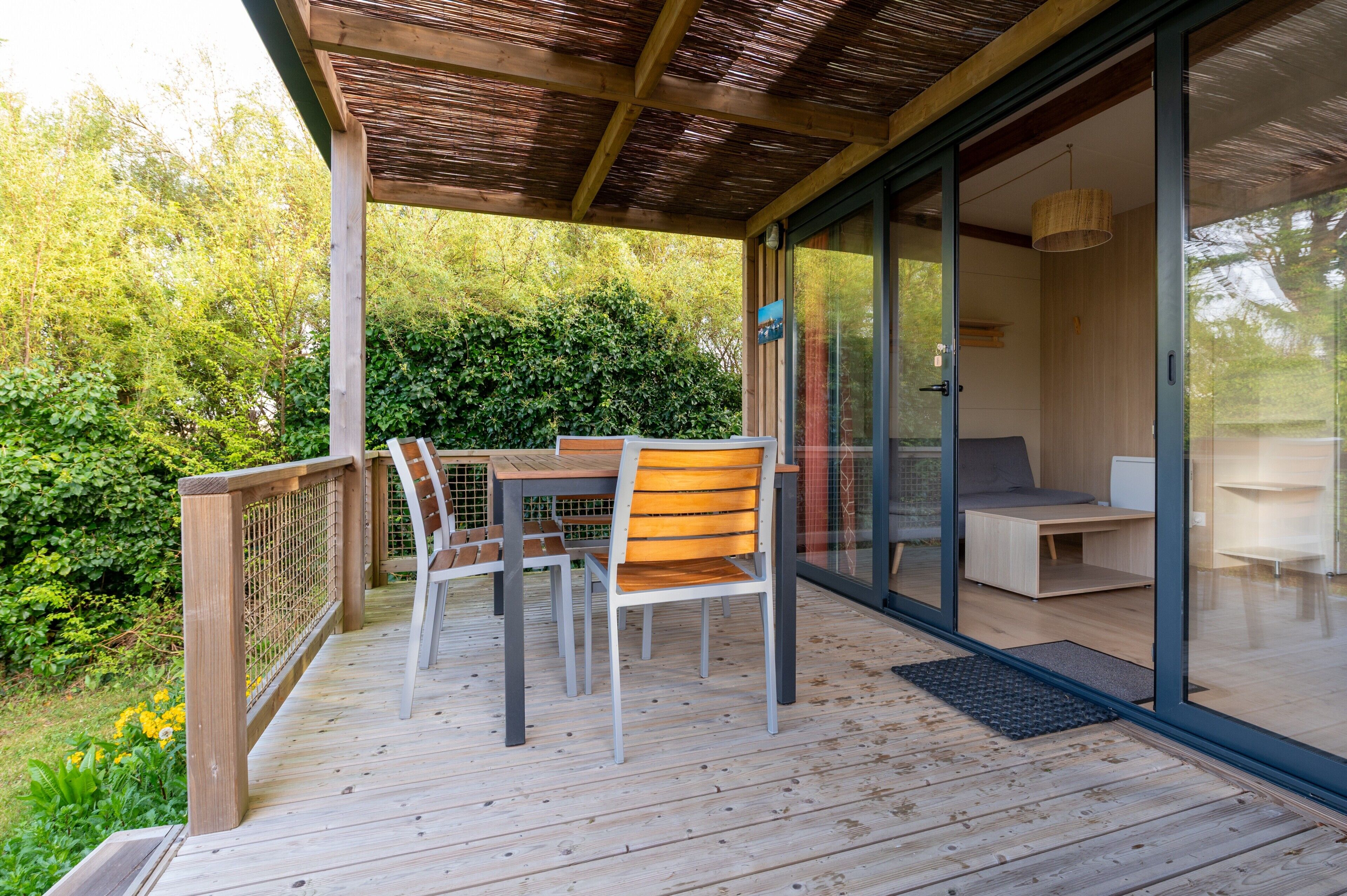 Een houten terras buiten met een tafel en stoelen, een glazen deur naar een kamer en uitzicht op groen.