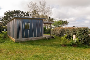A small, single-story building with a flat roof, a door, and a window, surrounded by a grassy area and dense shrubbery. - Chalet "30m² Le phare d'Eckmühl" in Plouhinec, 500m from the beach, sleeps 5 (Plouhinec)