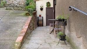 Un étroit sentier de pierres mène à un bâtiment rustique doté d’une table et de chaises en bois, de plantes en pot et d’une porte.