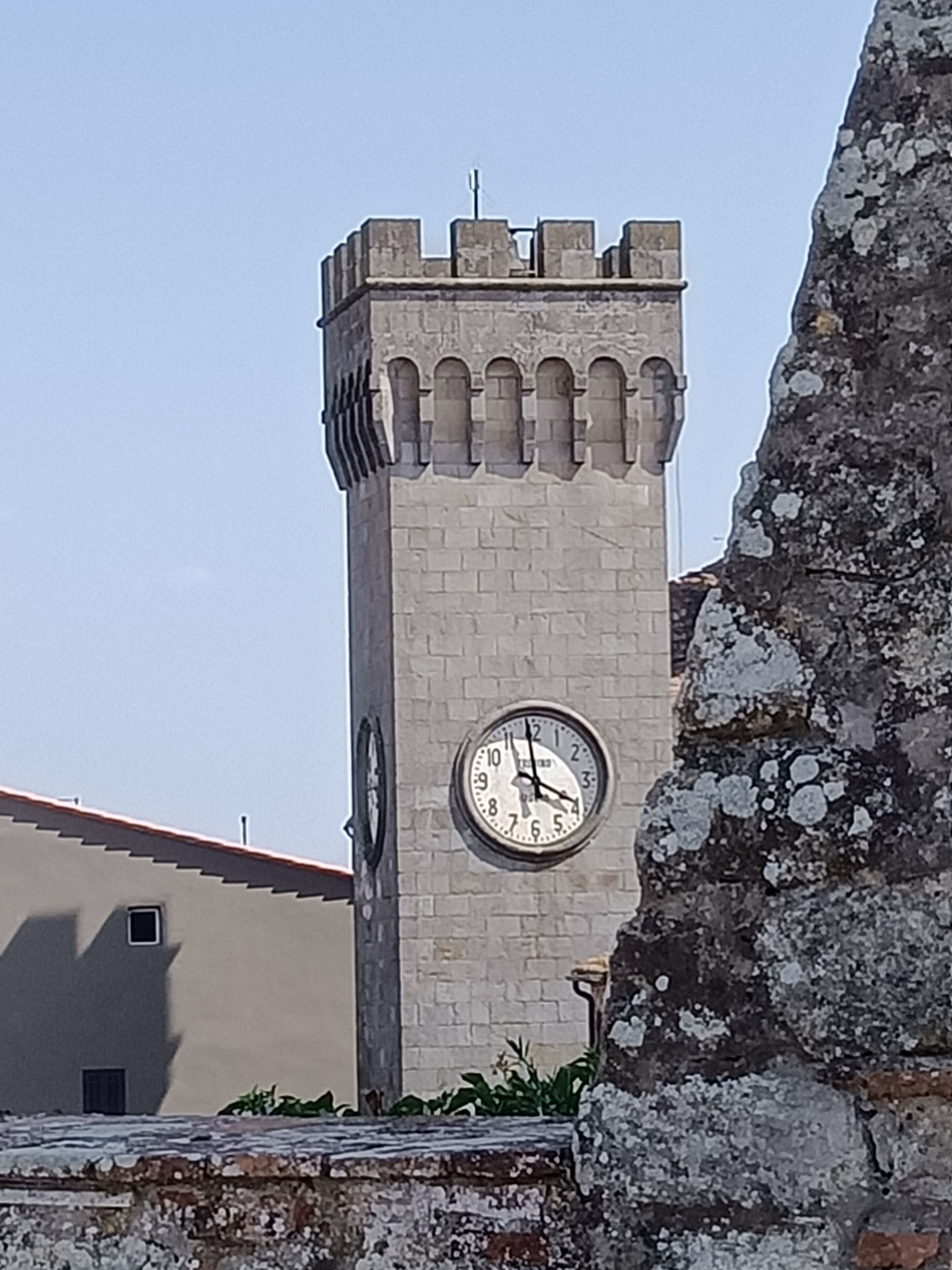 Uma torre do relógio com um mostrador redondo na lateral, uma parede de pedra e um edifício com telhado inclinado ao fundo.