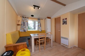 Ein Zimmer mit gelber Couch, einem Holzespeisetisch mit Stühlen, einem Fenster mit Vorhängen, einem Heizkörper und einer Tür.