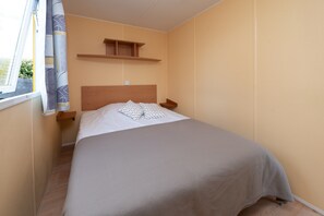 Una cama individual con cabecera de madera, una mesita de noche, una repisa sobre la cama y una ventana con cortinas.