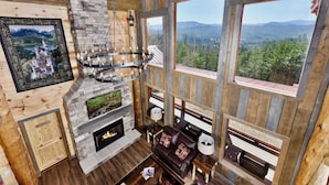 Interior acogedor de una cabaña con chimenea de piedra, paredes de madera y ventanas amplias con vistas a la montaña.