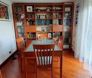 Une pièce avec une table et des chaises en bois, une bibliothèque remplie de livres, un tableau encadré et une lampe.