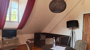 Una habitación acogedora con una mesa redonda, sillas de mimbre, un sofá, un televisor sobre un mueble bajo y una lámpara de pie.