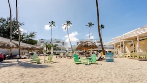 Een strand met rieten parasols, groene stoelen en een gebouw met een blauw dak op de achtergrond.
