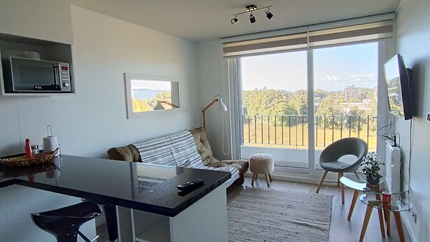 A modern living space with a kitchen area, a sofa, a dining table, and a balcony with a view. - Kasa and family 809 (Valdivia)