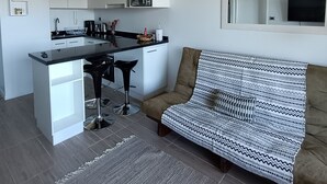 A modern kitchen with white cabinetry, a central island, and a dining area with a black bar stool. There is a sofa with a patterned throw blanket and a flat-screen TV mounted on the wall. - Kasa and family 809 (Valdivia)