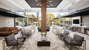 A spacious, modern lounge area with a central coffee table, comfortable seating, and large windows offering views of the outdoors. - Landing | Chic 1BD, Game Room, Courtyard (Birmingham)