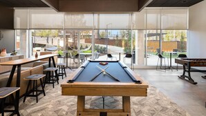 A pool table in a well-lit room with large windows, a bar area, and outdoor seating visible through the windows. - Landing | Chic 1BD, Game Room, Courtyard (Birmingham)