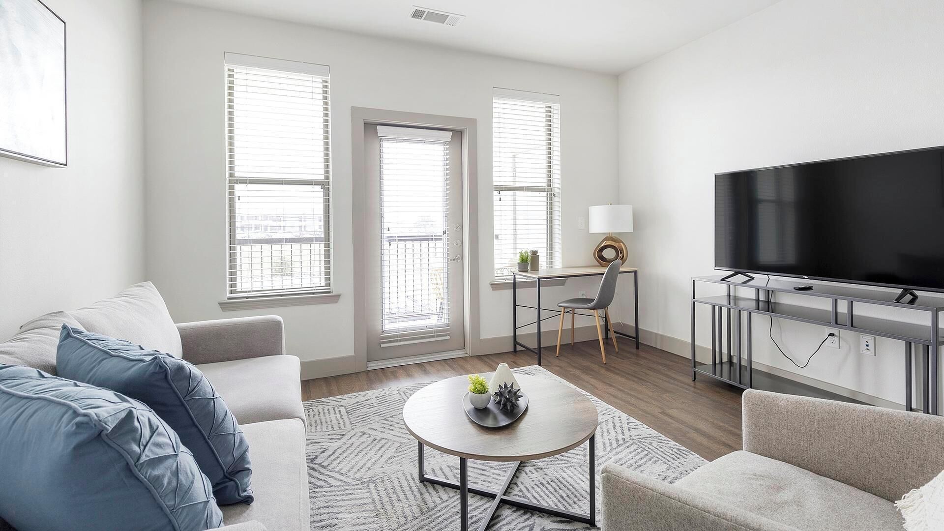 Ein modernes Wohnzimmer mit einer Couch, Sesseln, einem Couchtisch und einem Fernseher. Zwei Fenster mit Jalousien, eine Tür und ein Schreibtisch mit Lampe sind ebenfalls vorhanden.