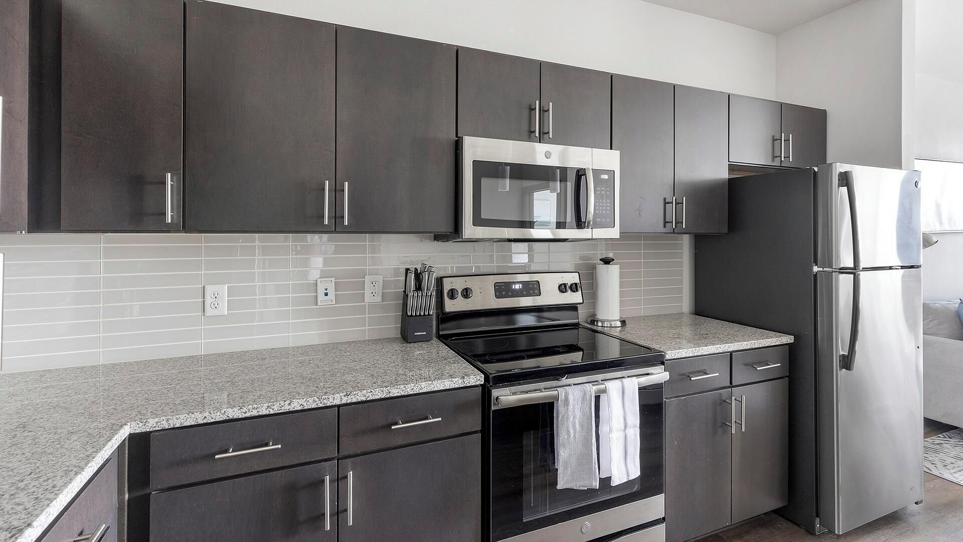 A modern kitchen with dark cabinets, a stainless steel refrigerator, a microwave, and a stove.