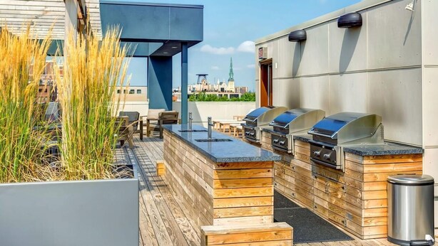 Area cucina esterna con diverse griglie barbecue, posti a sedere in legno e vista sullo skyline della città.