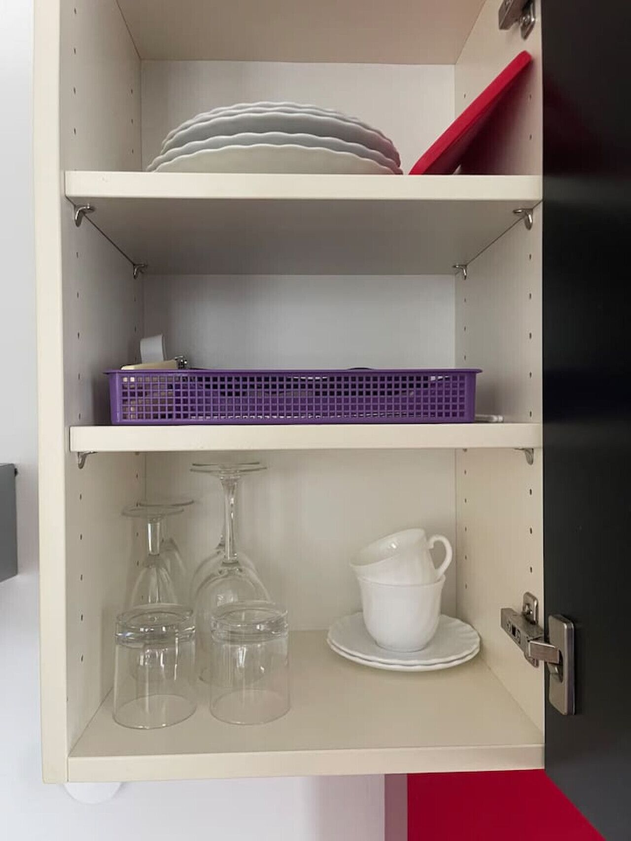 A kitchen cabinet with three shelves. The top shelf holds plates and a red tray. The middle shelf has a purple basket. The bottom shelf contains wine glasses and a white pitcher.