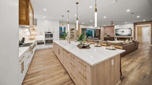 Cozinha moderna com armários brancos, uma ilha central e um espaço de jantar com sofá e televisão.