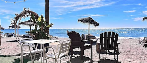 Un'area relax in spiaggia con tavoli e sedie, una palma e un ombrellone di paglia.