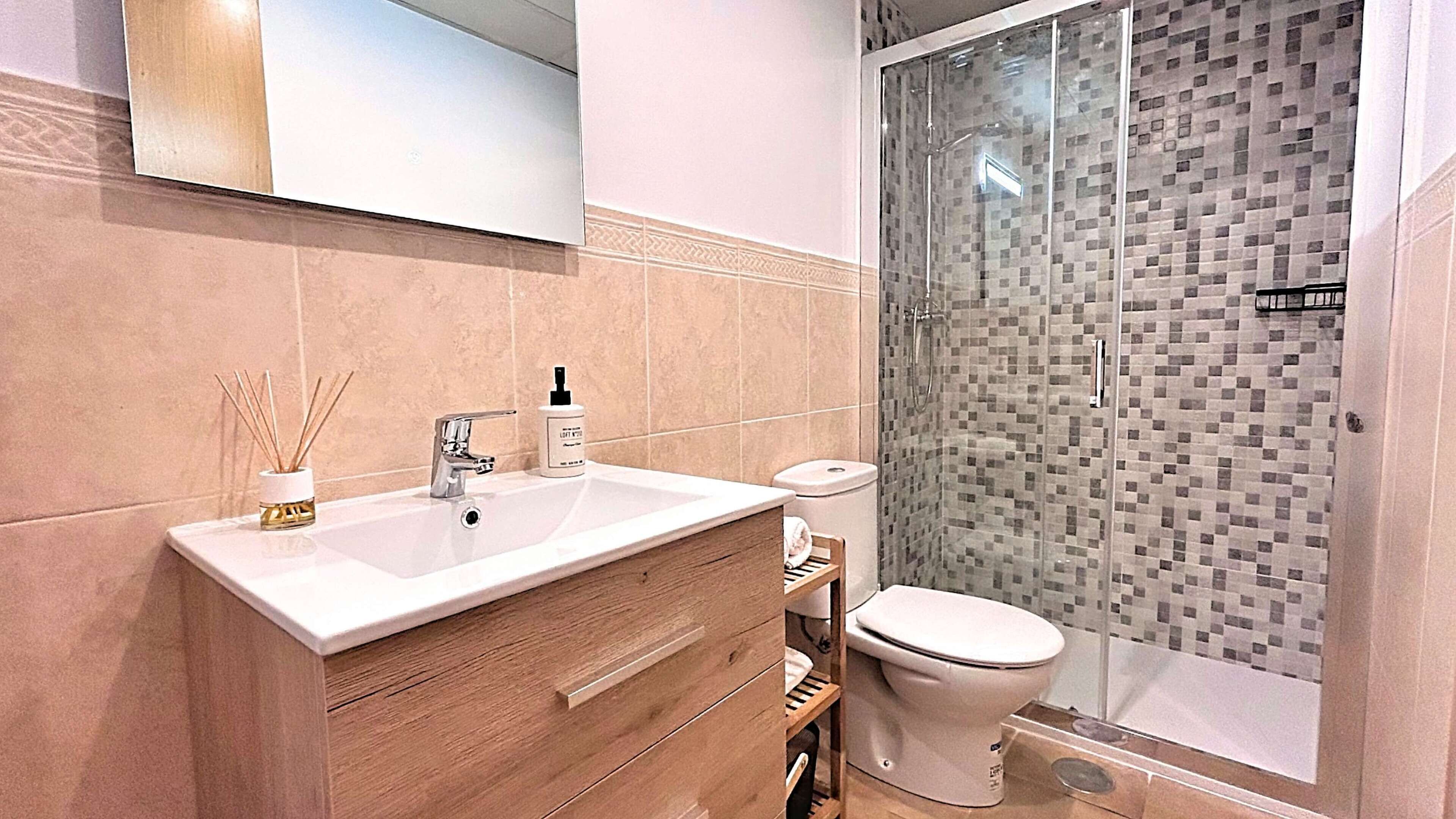 A modern bathroom with a white sink, wooden vanity, and a glass shower enclosure with mosaic tiles.