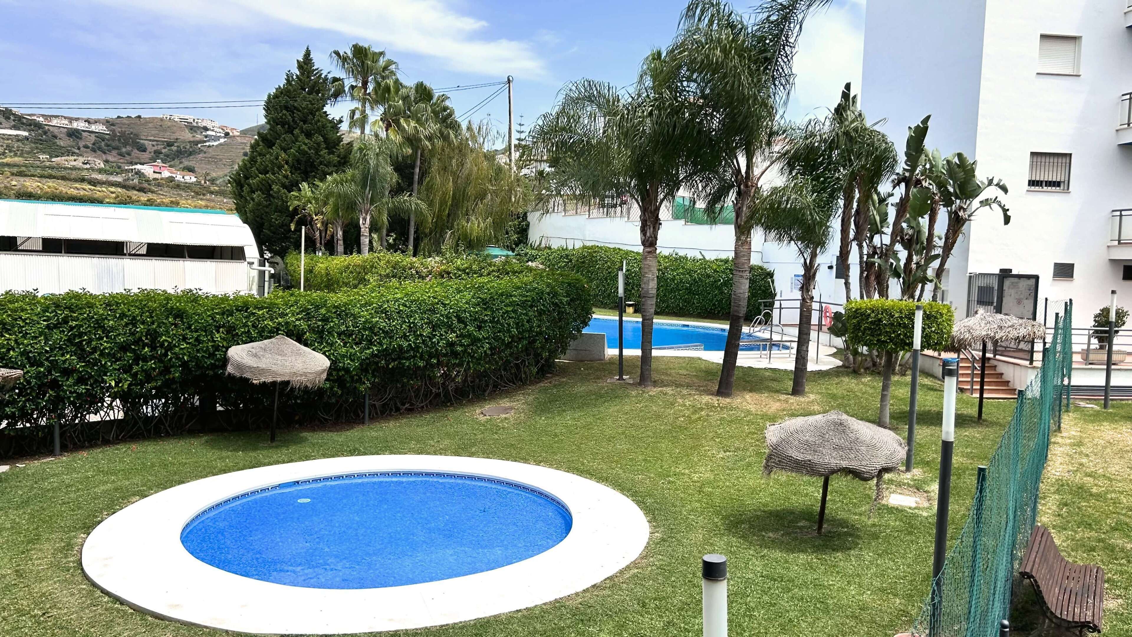 Une piscine entourée de pelouse, de palmiers et d’un bâtiment blanc, avec une autre piscine bleue en arrière-plan.