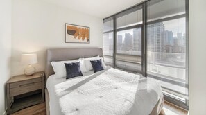Een slaapkamer met een bed, nachtkastje, lamp en raam met jaloezieën.