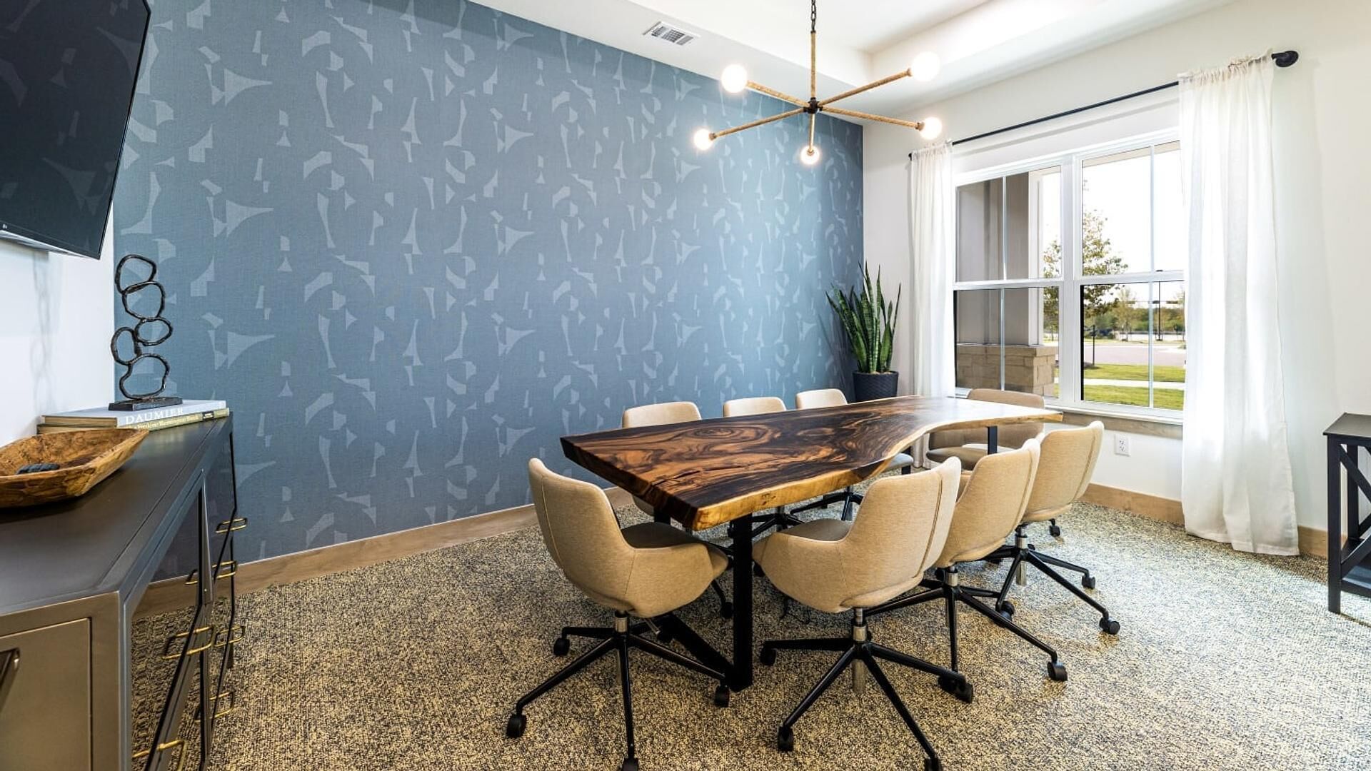 A conference room with a large wooden table, six chairs, a potted plant, and a window with curtains.